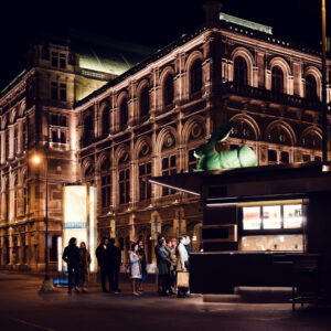 Long line of people in front of Bitzinger Würstelstand, with the historic State Opera in the background.