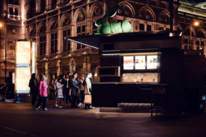 Excited crowd at Bitzinger Würstelstand, with Vienna's cultural landmarks in the background.