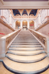 The grand staircase inside Albertina Modern museum, leading up to arched doorways and a beautifully decorated ceiling with blue and gold detailing.