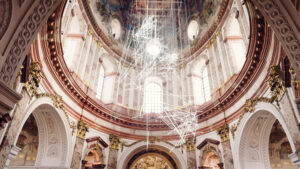A contemporary art installation at St. Charles Church (Karlskirche), resembling a delicate web of light, hangs from the ceiling, creating a striking contrast between the baroque architecture and modern art.