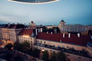 Evening view from MQ Libelle terrace overlooking illuminated Museumsquartier Vienna with golden hour lighting and historic architecture.