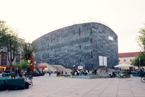 MUMOK Vienna's distinctive dark monolithic form viewed from the MuseumsQuartier main plaza with visitors enjoying the cultural surroundings.