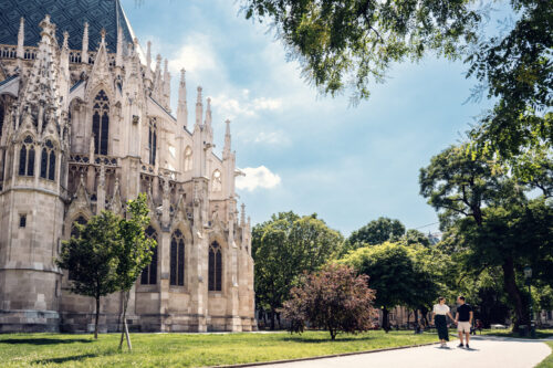 Adriana and Mario strolling near Votivkirche Vienna, with the church's detailed limestone facade and pointed arches creating a dramatic backdrop.