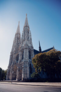 Front facade of Votivkirche Vienna with intricate stone carvings, rose window, and Gothic arches illuminated by summer afternoon sunlight.
