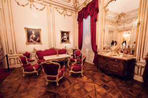 Baroque conference room at Schönbrunn Palace (Schloss Schönbrunn) with red velvet curtains and period furniture, imperial Habsburg palace interior featuring ornate wooden chairs around antique table, golden rococo wall decorations and portrait paintings, crystal chandelier and parquet flooring.