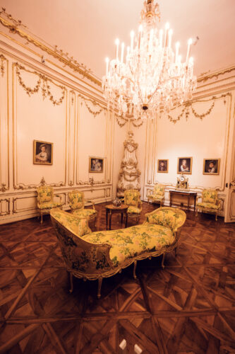 Luxurious palace chamber in Schönbrunn Palace (Schloss Schönbrunn) with gilded rococo wall panels and historic furniture, baroque Austrian imperial interior with yellow period chairs and wooden table, ornate golden decorations and crystal lighting.
