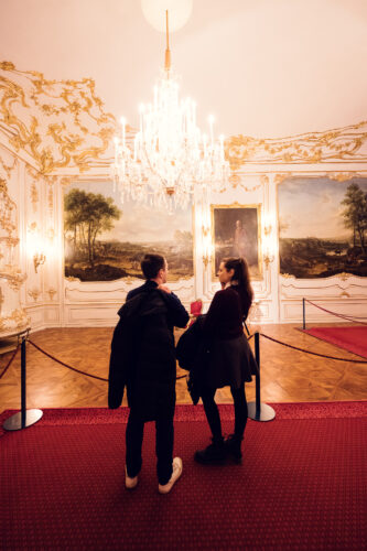 Adriana und Mario betrachten Kunstwerke im Schloss Schönbrunn, barocke kaiserliche Kunstsammlung mit historischen Gemälden und prunkvollen goldenen Rahmen.