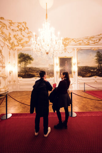 Adriana and Mario viewing artwork at Schönbrunn Palace (Schloss Schönbrunn), baroque imperial palace art collection with historic paintings and ornate golden frames.