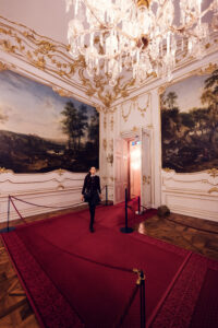 Adriana on red carpet at Schönbrunn Palace (Schloss Schönbrunn), baroque imperial palace interior with ornate golden decorations and crystal chandeliers.