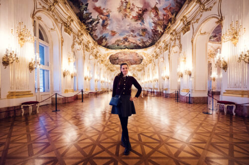 Adriana in elegant pose at Schönbrunn Palace (Schloss Schönbrunn), baroque Austrian imperial palace with elaborate golden ornaments and period furniture.