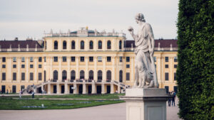 Baroque garden statue Schönbrunn with ornate stone sculpture and yellow Schönbrunn Palace facade visible in background through formal gardens.