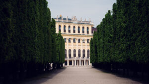 Schönbrunn Palace side view through tree-lined green pathway with mature foliage framing yellow baroque palace facade.