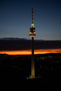 Vienna's Donauturm illuminated at night, its 252-meter structure glowing with LED lights, featuring the rotating observation deck and restaurant levels visible against the dark sky.