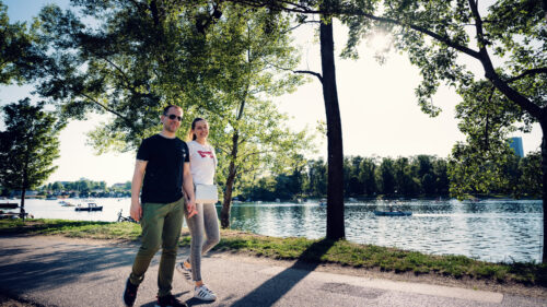 Adriana and Mario walking along Alte Donau riverbank in Vienna with natural waterfront landscape and calm water in background.