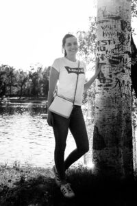 Porträt von Adriana an einem Baum am Ufer der Alten Donau in Wien mit ruhiger Wasseroberfläche und Uferbewuchs dahinter.