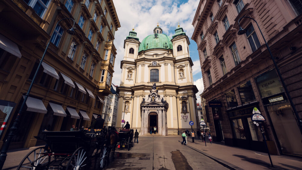 Street-level view of St. Peter's Church Vienna (Peterskirche) showing the elliptical structure, twin bell towers, and the ornate entrance portal with stone sculptures representing the church's historical significance.