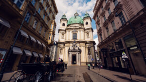 Street-level view of St. Peter's Church Vienna (Peterskirche) showing the elliptical structure, twin bell towers, and the ornate entrance portal with stone sculptures representing the church's historical significance.