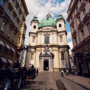 Street-level view of St. Peter's Church Vienna (Peterskirche) showing the elliptical structure, twin bell towers, and the ornate entrance portal with stone sculptures representing the church's historical significance.