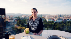 Adriana drinking cocktail at Aurora Rooftop Bar Vienna, modern rooftop terrace atmosphere with Vienna skyline backdrop and contemporary bar setting.