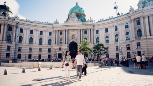 Adriana and Mario walking hand in hand across Michaelerplatz with the Hofburg Palace's Michaelertrakt dome in the background.