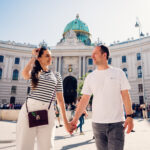Adriana und Mario gehen gemeinsam am Michaelerplatz in Wien mit der verzierten Barockfassade der Hofburg entlang.