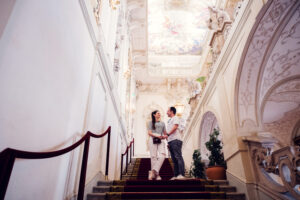 Adriana and Mario posing in Palais Kinsky Vienna baroque interior hall with ornate ceiling frescoes and classical palace decorations.