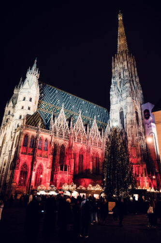 Illuminated St. Stephen's Cathedral (Stephansdom) with Christmas market vendors on Stephansplatz, Vienna winter evening.
