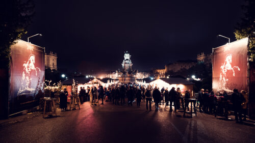 Rote Banner mit einem steigenden weißen Pferd rahmen nachts eine belebte Gasse zum Weihnachtsmarkt auf dem Maria-Theresien-Platz ein.