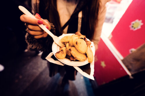 Am Weihnachtsmarkt setzt eine Hand mit roten Nägeln eine Gabel auf knusprige Kartoffelscheiben in einem Pappteller.