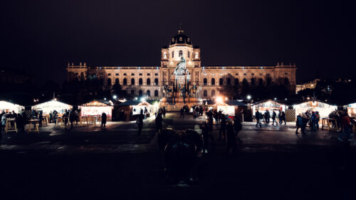 Hütten und Stehtische umringen den Weihnachtsmarkt auf dem Maria-Theresien-Platz, dahinter ragen das angestrahlte Denkmal und das Museum auf.