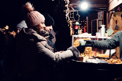 Lächelnd nimmt Adriana an einem Weihnachtsmarktstand mit Glühbirnen und Spiralkartoffeln eine Spitztüte aus Papier entgegen, Mario hinter ihr.