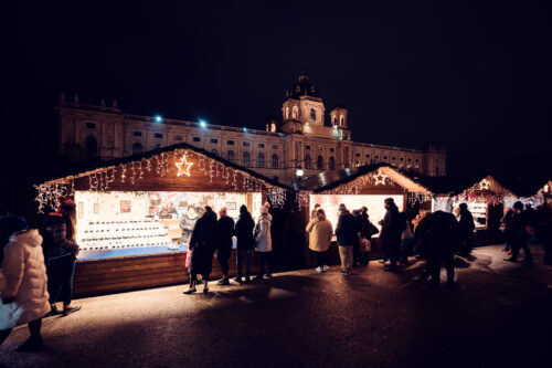 Aus Weihnachtsmarkthütten mit Sternen auf dem Dach fällt warmes Licht über den Maria-Theresien-Platz, dahinter die angestrahlte Museumskuppel.