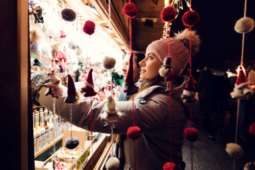 Adriana blickt an einem hell erleuchteten Weihnachtsmarktstand durch baumelnde rote Bommeln und Wichtelanhänger nach oben.