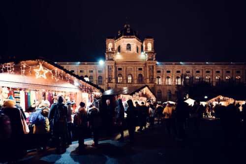 Bunte Strickschals leuchten unter Sternlichtern, während nachts viele Menschen über den Weihnachtsmarkt auf dem Maria-Theresien-Platz bummeln.