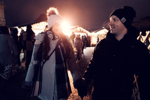 Adriana und Mario schlendern lächelnd Hand in Hand über den Weihnachtsmarkt.