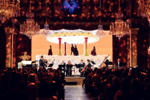 Orchestra performing on stage with LED backdrop and crystal chandeliers in the Strauss Hall.