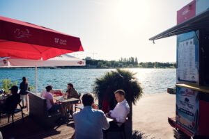 Guests at a Copa Beach bar terrace under red parasols beside the Neue Donau.