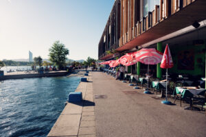Paved Copa Beach promenade lined with red parasols and restaurant terraces along the Neue Donau.
