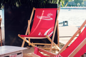 A red Copa Beach deck chair set on the sand by the water.