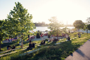 People relaxing on the grassy Copa Beach lawn above the Neue Donau at golden hour.