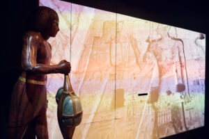 Egyptian figure holding a scarab in front of a wall projection of ancient hieroglyphics.
