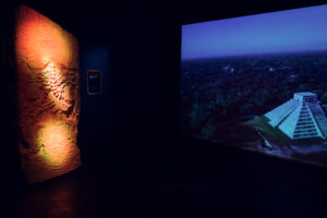 Illuminated Maya stone relief next to a screen showing an aerial view of the Kukulcan pyramid.