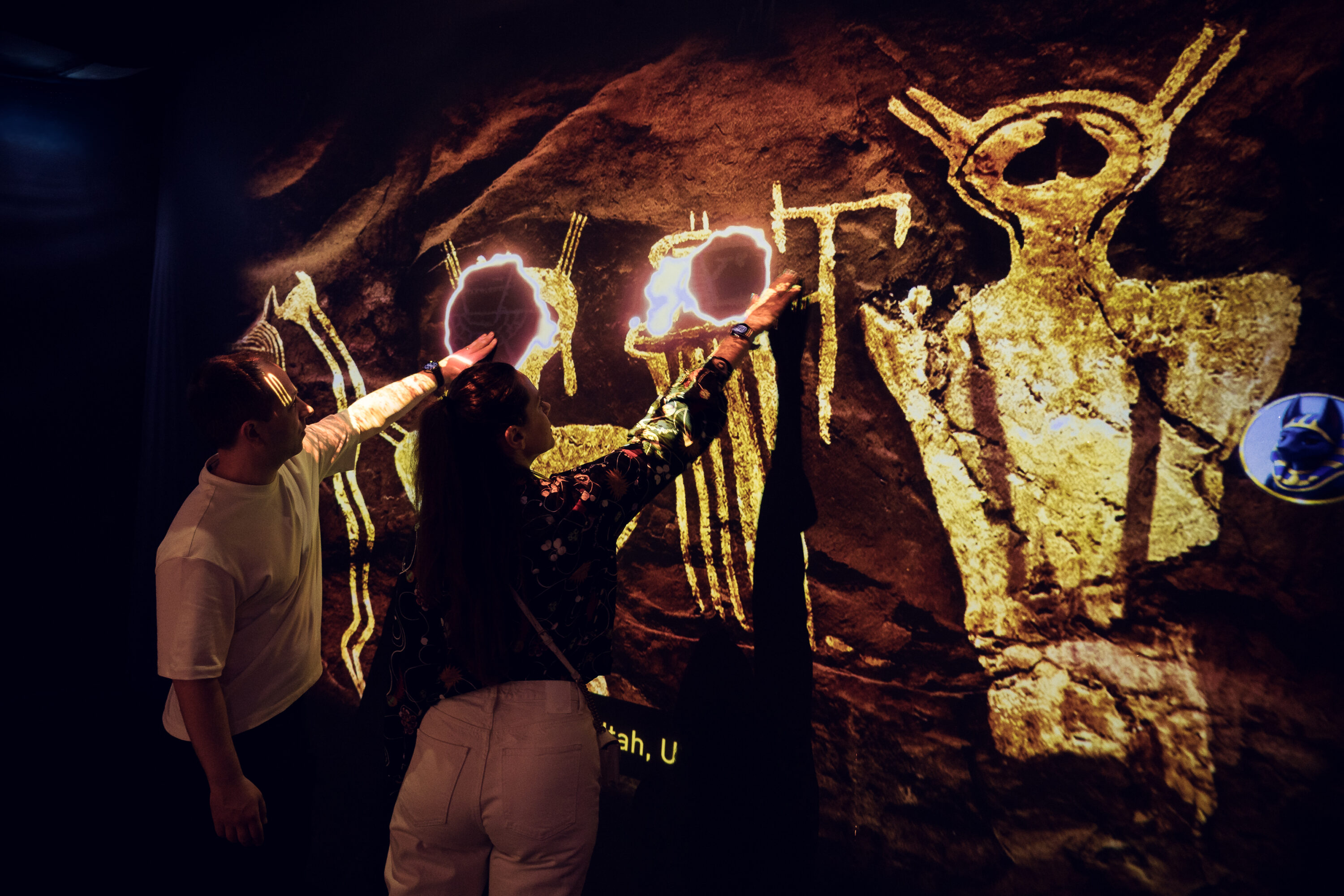 Adriana and Mario touching an interactive wall projection of glowing cave paintings and ancient symbols.