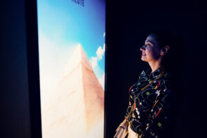 Adriana standing in profile beside a vertical screen showing the Great Pyramid of Giza.