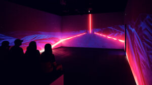 Visitors seated in the 360-degree projection room watching a landscape scene with a pink neon light strip along the walls.