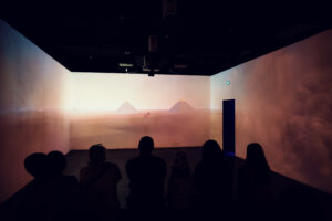 Visitors in the 360-degree room watching a desert sunset projection with distant pyramids on the horizon.