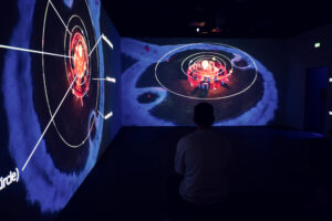 Visitor sitting on the floor watching circular orbital projections on the walls of a dark room.