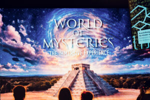 Large projection screen showing the World of Mysteries title card with a Maya pyramid and starry sky.
