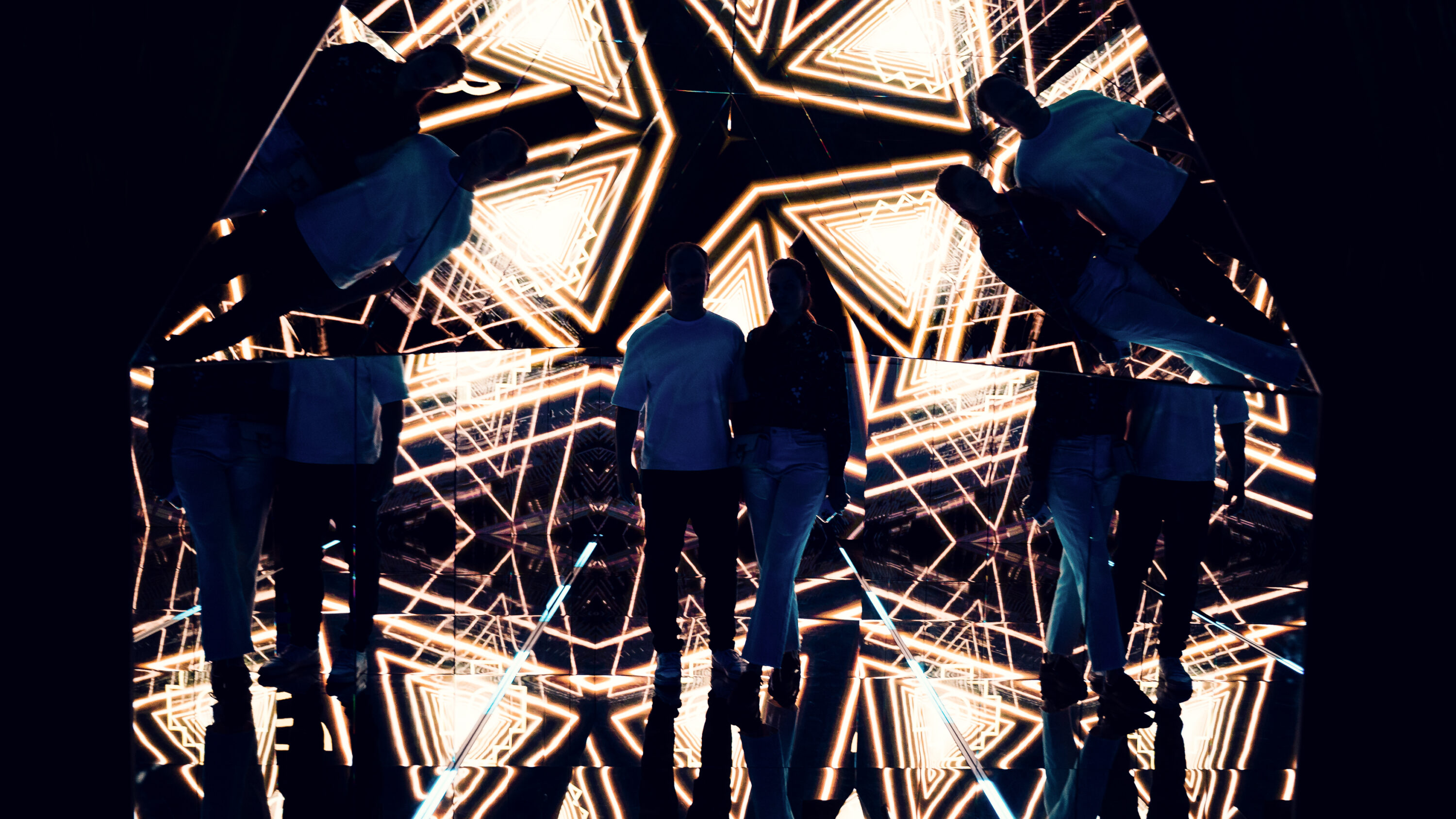 Adriana and Mario standing in the pyramid room with geometric golden projections reflected around them.