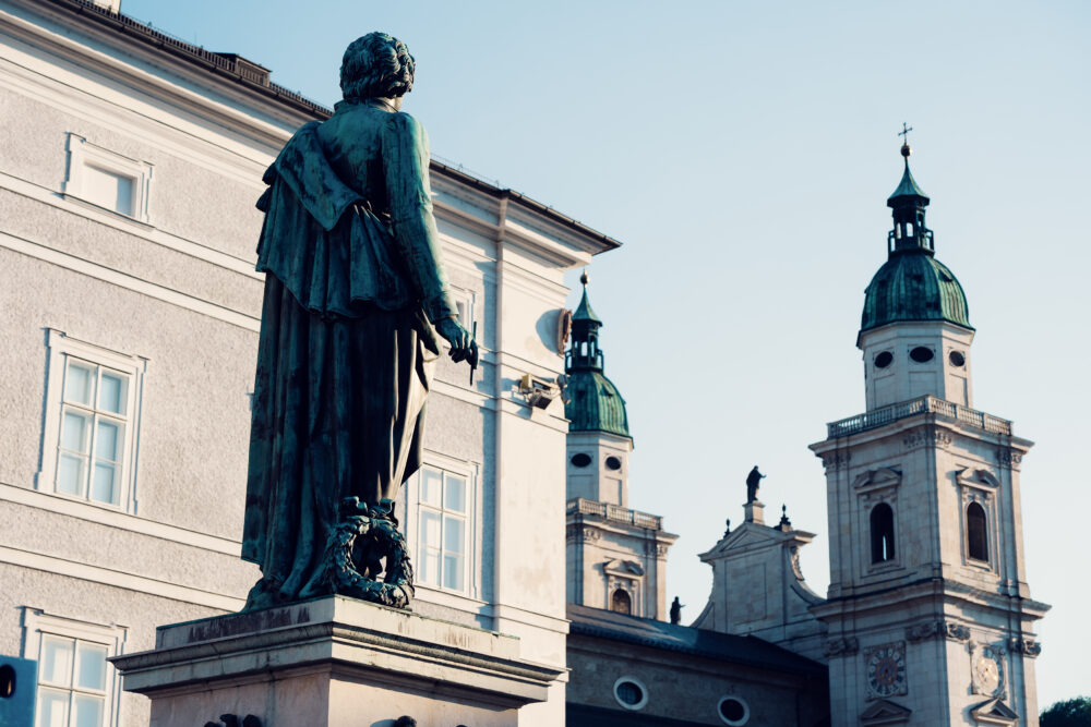 The cathedral's two towers with green domes rise beside the Mozart statue at Mozartplatz under a clear sky.